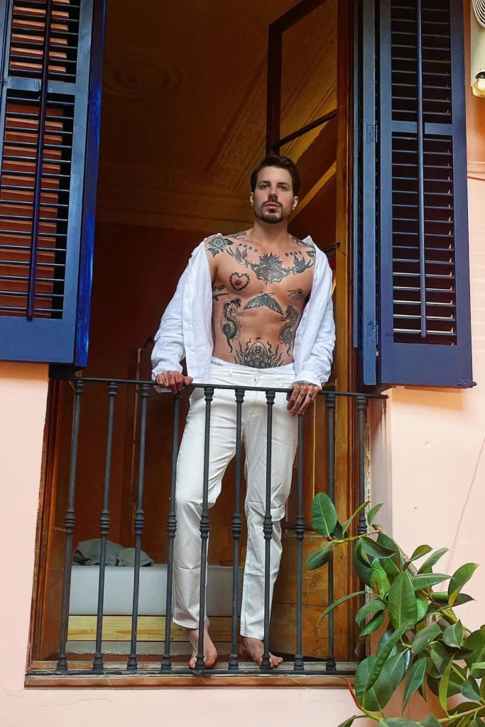 Enea, masajista de Bliss Barcelona, posando en un balcón con camisa blanca abierta y pantalón blanco, estilo mediterráneo